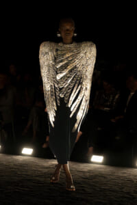 Model walks a runway in a dramatic silver dress with wing-like metallic panels, audience in the dark background.