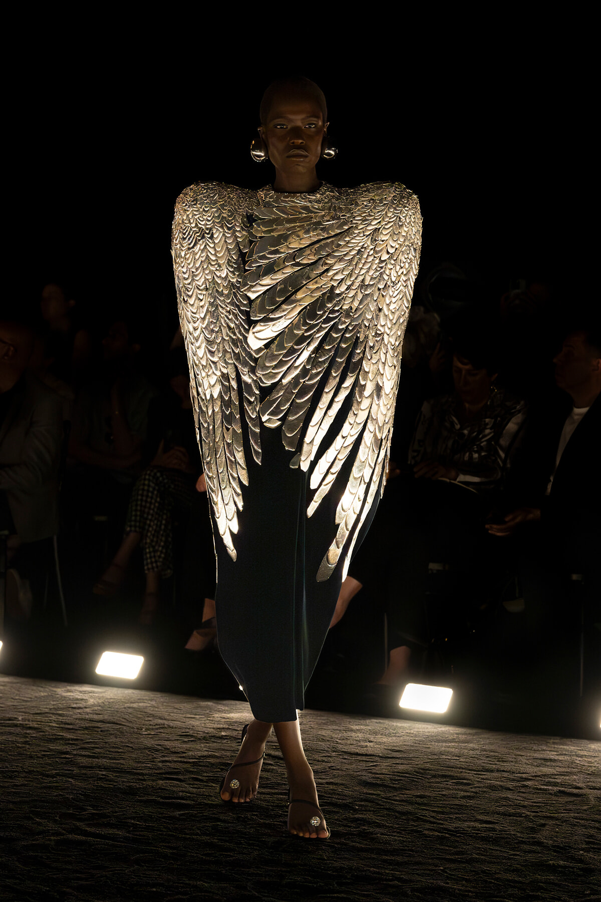 Model walks a runway in a dramatic silver dress with wing-like metallic panels, audience in the dark background.