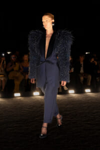 Model walks a fashion runway in a navy suit with a dramatic beaded feathered jacket and statement heels under spotlights.