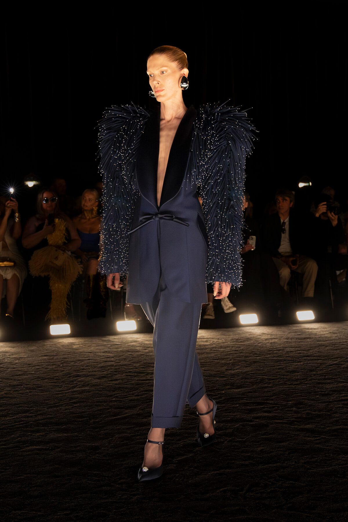 Model walks a fashion runway in a navy suit with a dramatic beaded feathered jacket and statement heels under spotlights.
