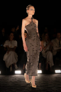 Model on a runway wearing a gray halter dress with triangular cutouts and spikes, rose choker, nude heels.