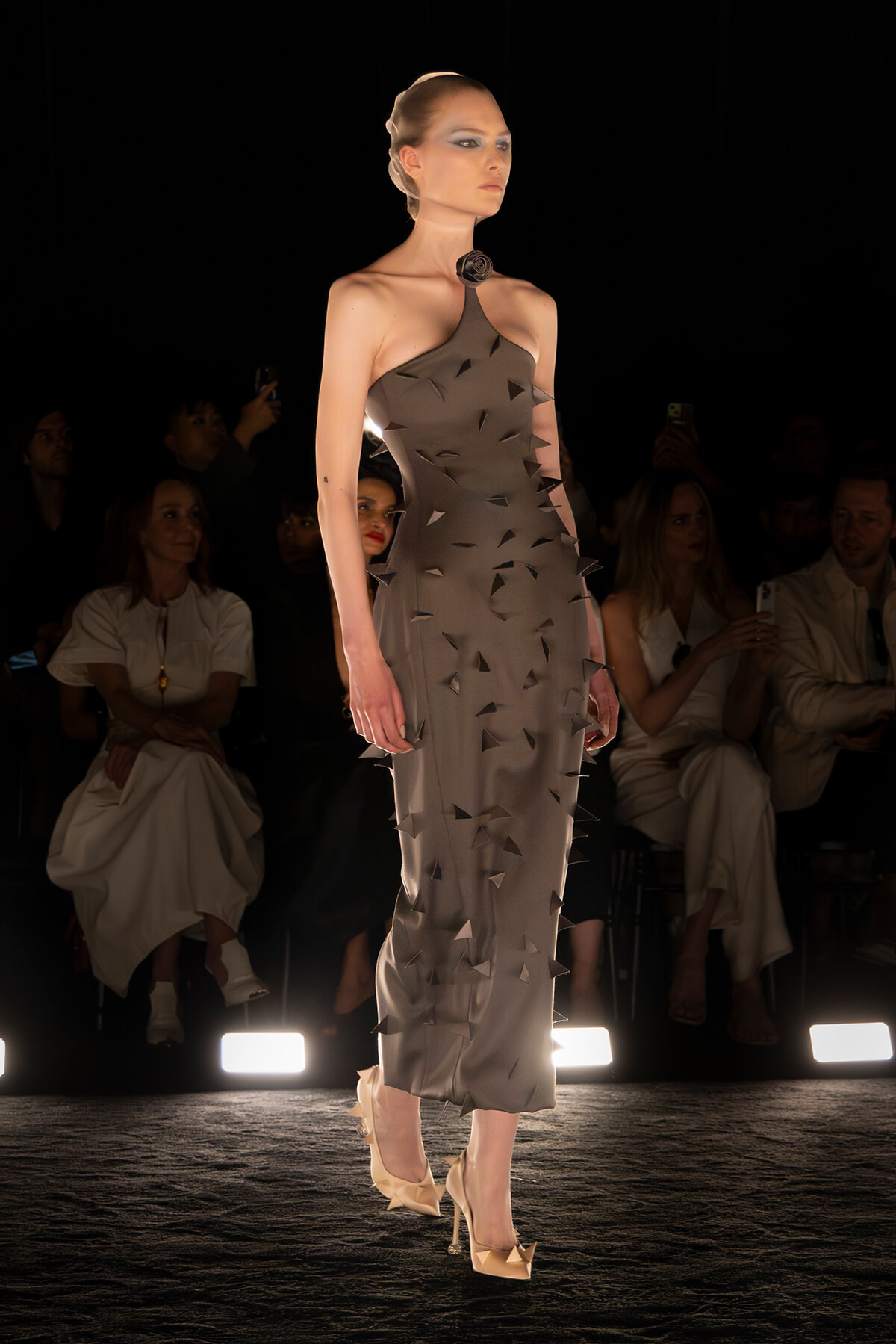 Model on a runway wearing a gray halter dress with triangular cutouts and spikes, rose choker, nude heels.