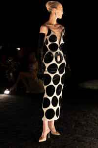 Model on a runway wearing a black-and-white polka-dot gown with circular cutouts, long black gloves, and beige heels.