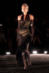 Model on a runway wearing a metallic halter gown with diagonal draping and a long fringed train, dark backdrop and audience in the background.] ,