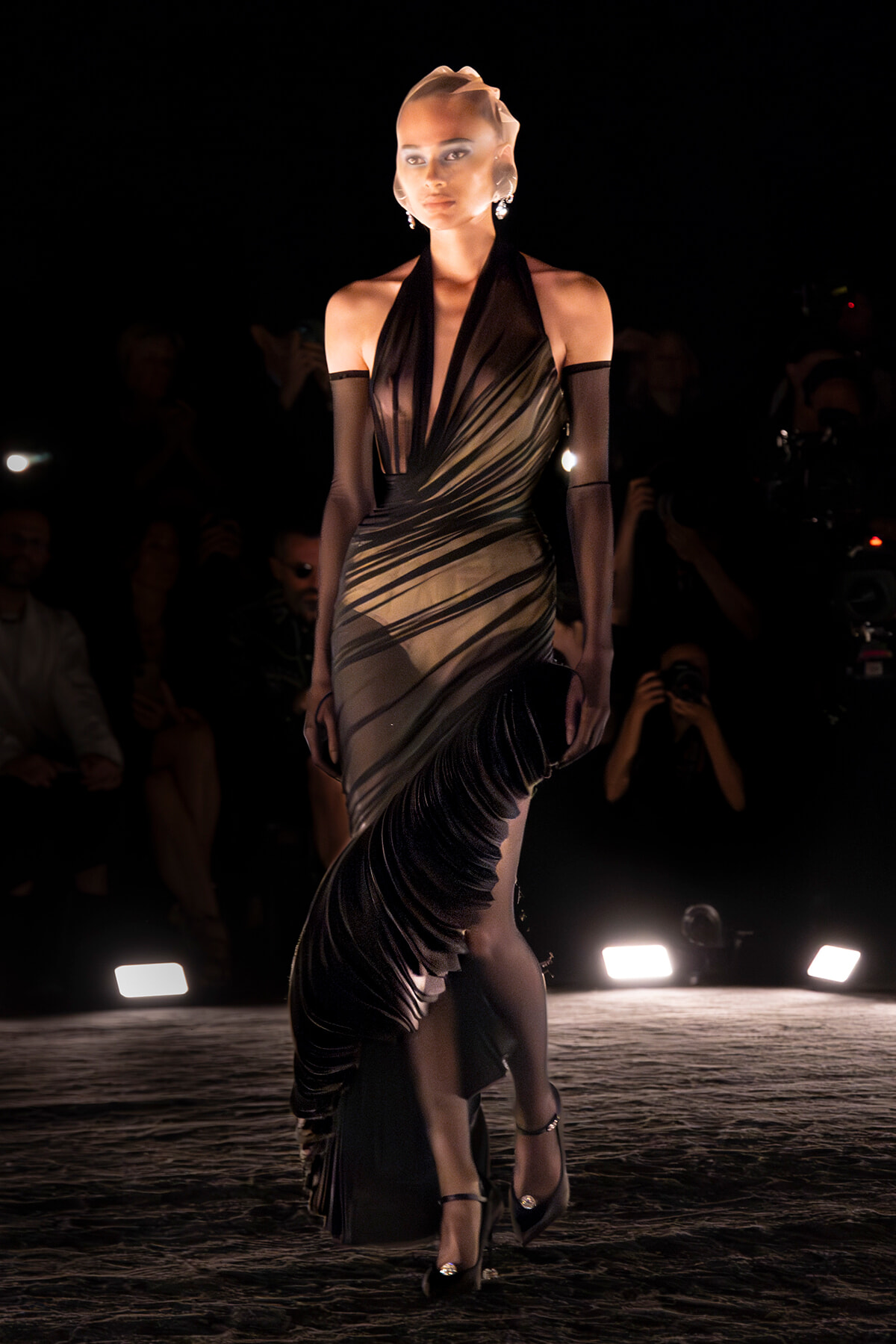 Model on a runway wearing a metallic halter gown with diagonal draping and a long fringed train, dark backdrop and audience in the background.] ,
