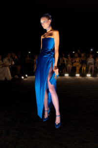 Model in a cobalt blue strapless gown with a high slit walks a runway, clutching a matching blue clutch as spectators watch in the dark.
