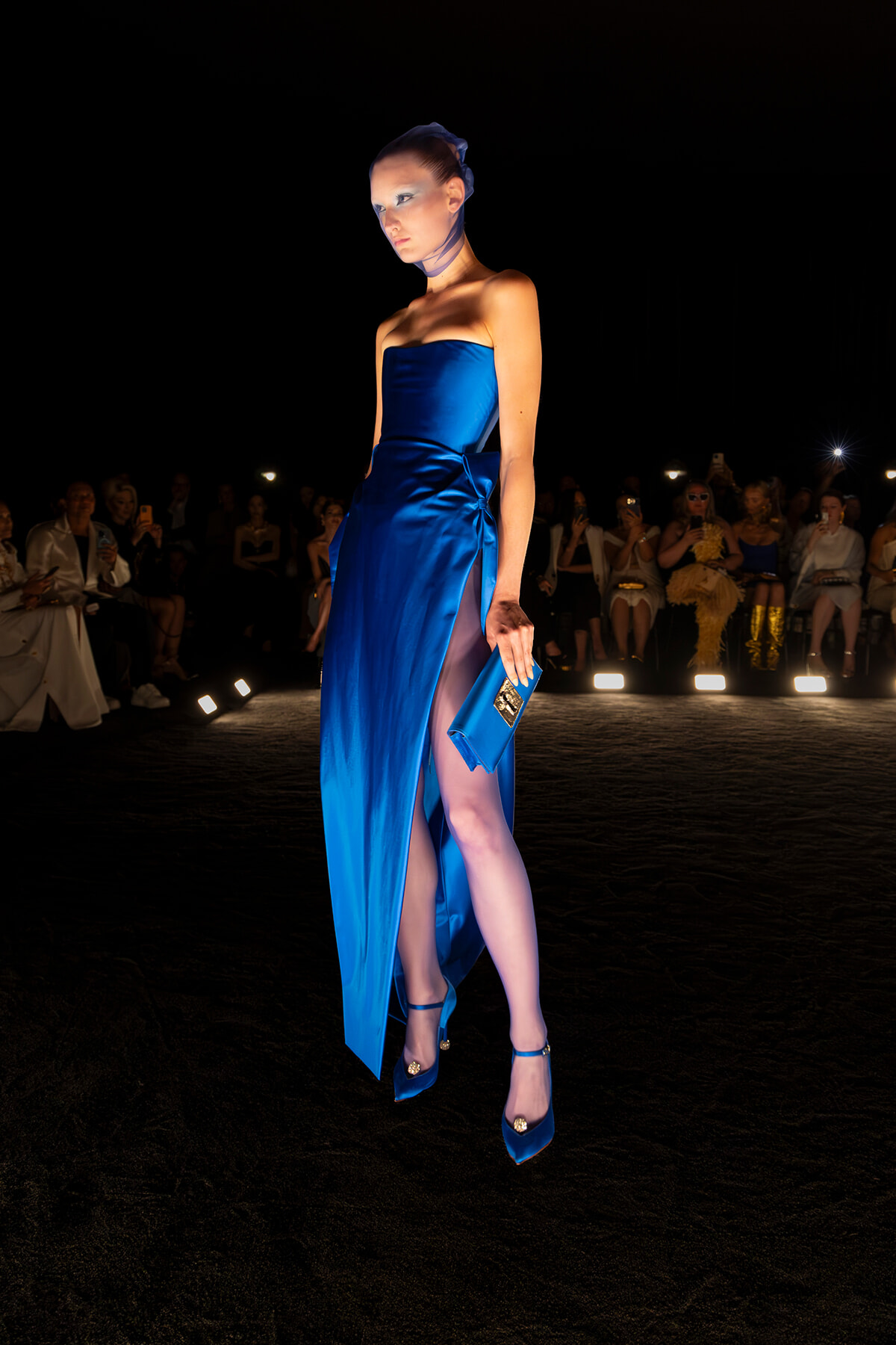 Model in a cobalt blue strapless gown with a high slit walks a runway, clutching a matching blue clutch as spectators watch in the dark.