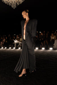 Model on a dark runway in a long black draped dress with open front, exposing midriff; chandelier overhead and audience in the background.