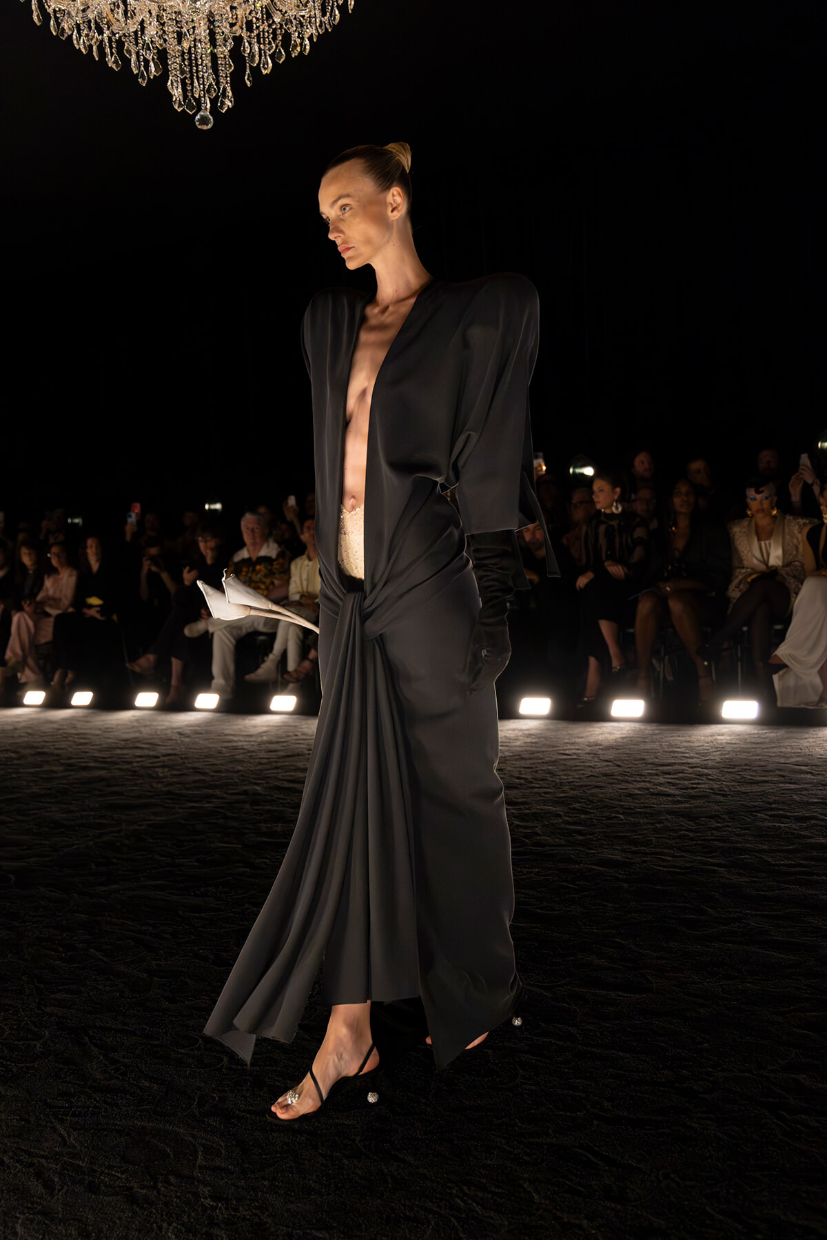 Model on a dark runway in a long black draped dress with open front, exposing midriff; chandelier overhead and audience in the background.
