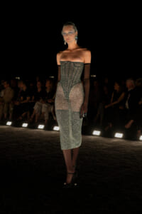 Model on a dim runway wearing a shimmering strapless midi dress with sheer sides and long black gloves.