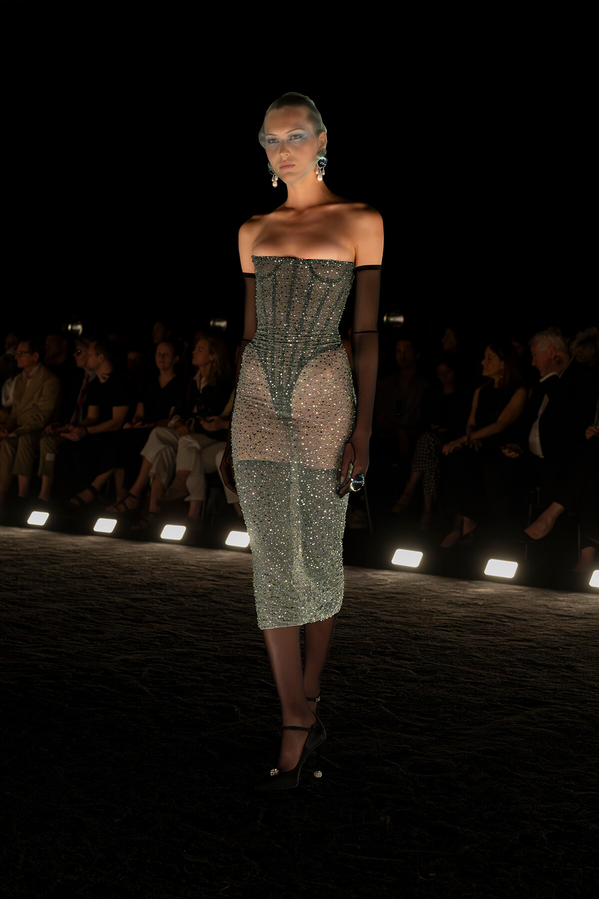 Model on a dim runway wearing a shimmering strapless midi dress with sheer sides and long black gloves.