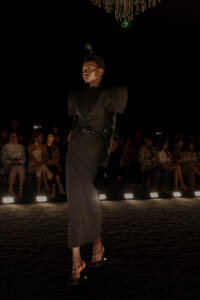 Model walks a dark runway in a dramatic black dress with structured shoulders, long gloves, and heels, audience watching in the background.