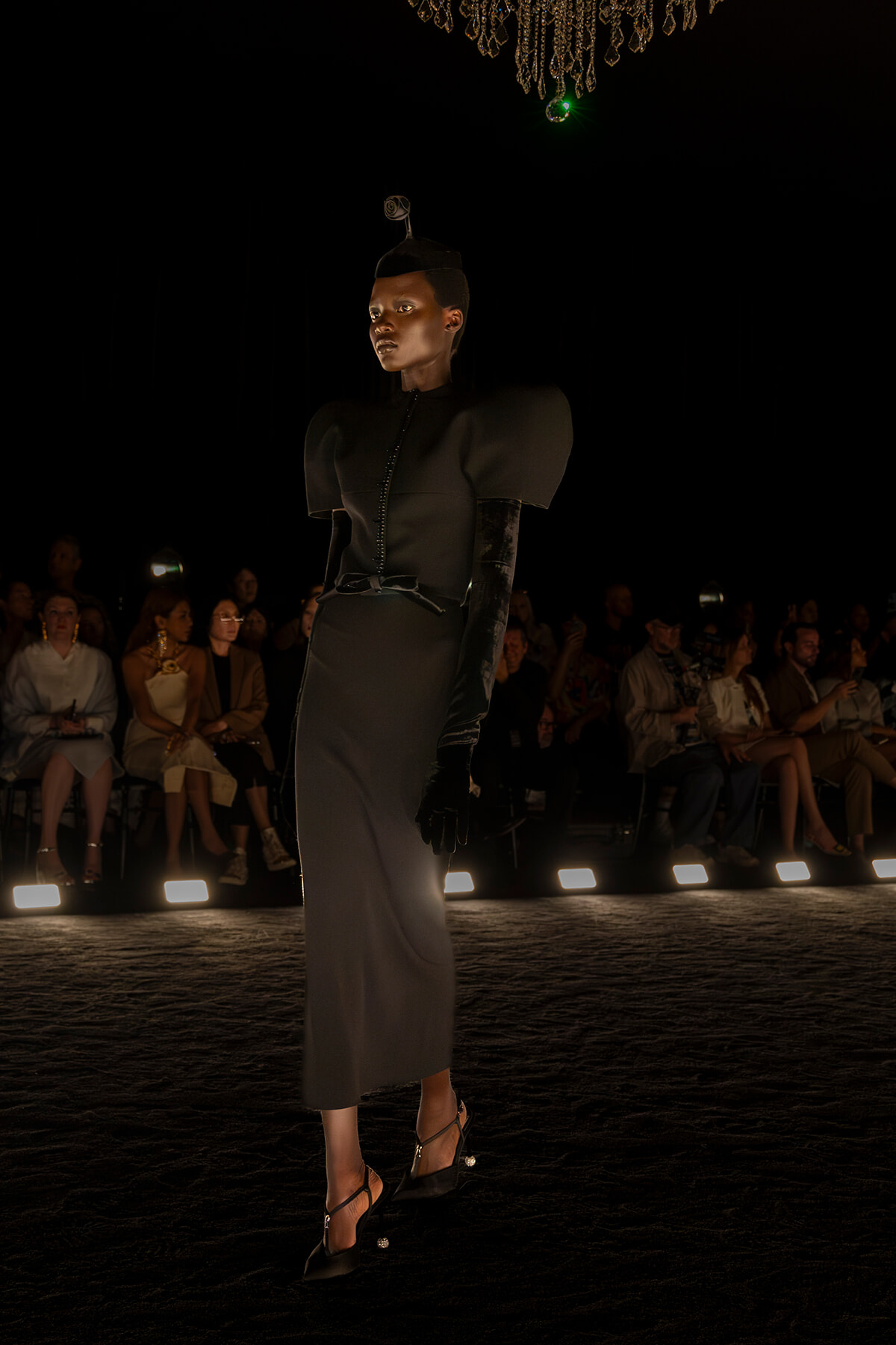 Model walks a dark runway in a dramatic black dress with structured shoulders, long gloves, and heels, audience watching in the background.