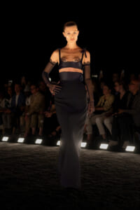 Model walks the runway in a black satin two-piece lingerie-inspired outfit with lace trim and sheer gloves, audience in background.
