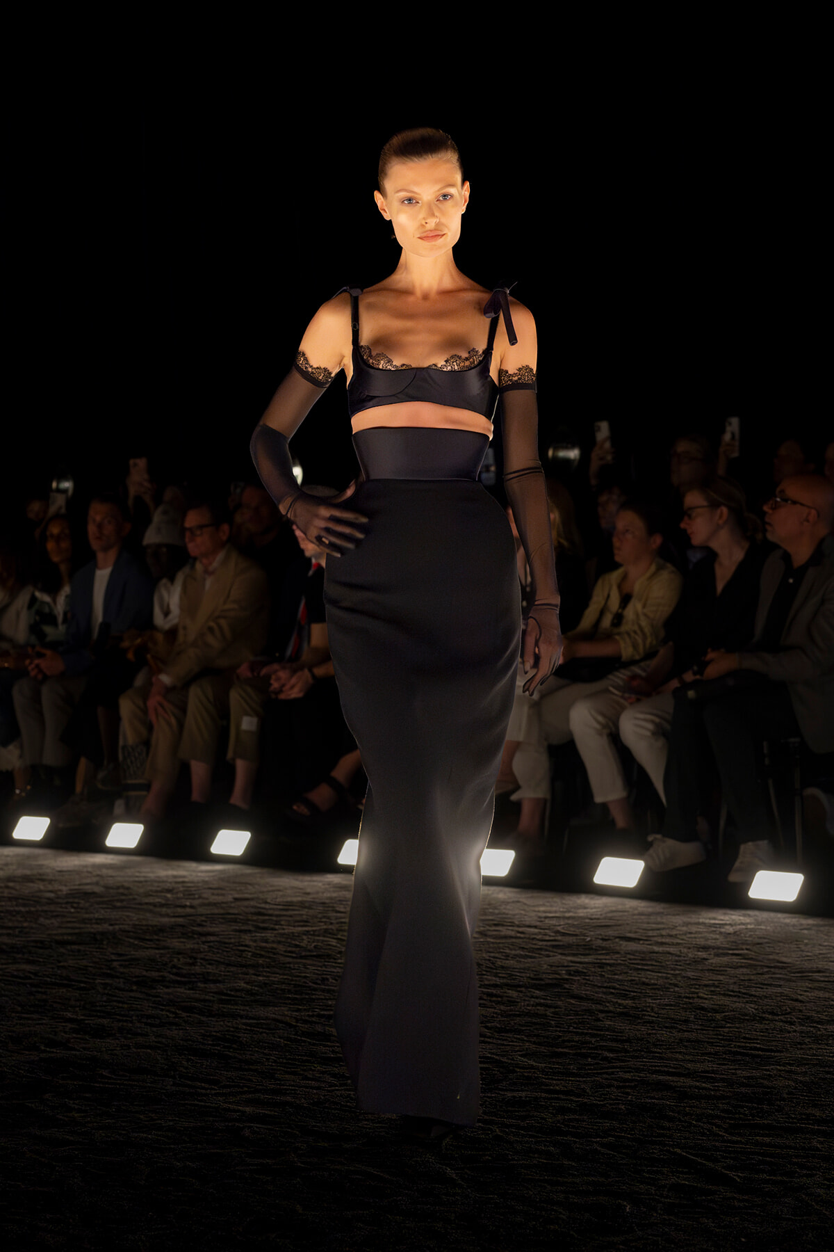 Model walks the runway in a black satin two-piece lingerie-inspired outfit with lace trim and sheer gloves, audience in background.
