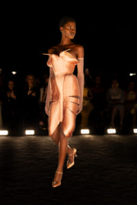 Model walks a fashion runway in a peach-toned sculptural dress with an off-shoulder bodice and sheer gloves, under spotlight.