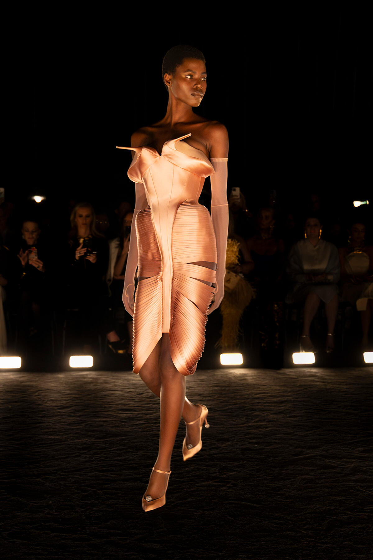 Model walks a fashion runway in a peach-toned sculptural dress with an off-shoulder bodice and sheer gloves, under spotlight.