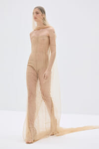 Model wearing a nude-toned, off-the-shoulder mesh gown with a long sleeve and sheer veil, standing in a studio.