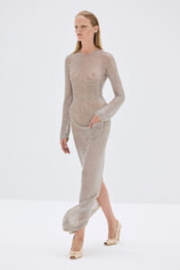 Pregnant model wearing a sheer, beaded beige knit dress with pockets and beige heels, with oxygen tubing around the nose.