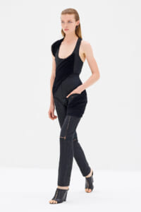 Model in a black textured jumpsuit with ruched wrap detail, standing against a white studio backdrop wearing open-toe heeled sandals