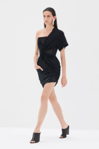 Model wearing a black one-shoulder draped dress with mesh panels and ruched detailing, walking in a white studio space with open-toe heeled sandals.
