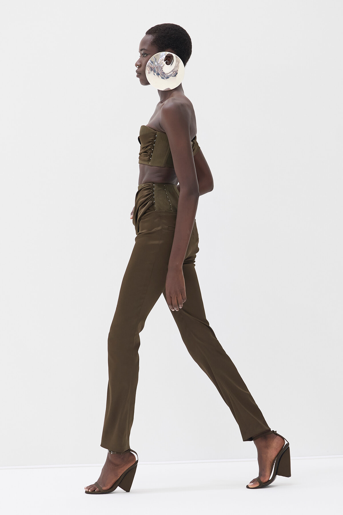 Profile view of a model walking in an olive-green strapless top and matching high-waisted pants, with a large circular metallic earring.