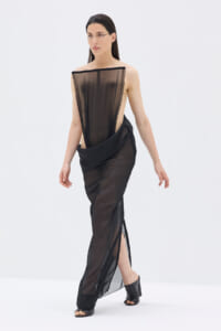 Model wearing a black sheer evening gown with a draped overlay, one-shoulder neckline, and a side slit.