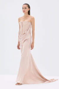Model wearing a blush pink strapless gown with a corset-style bodice and draped sash, floor-length with a long train.
