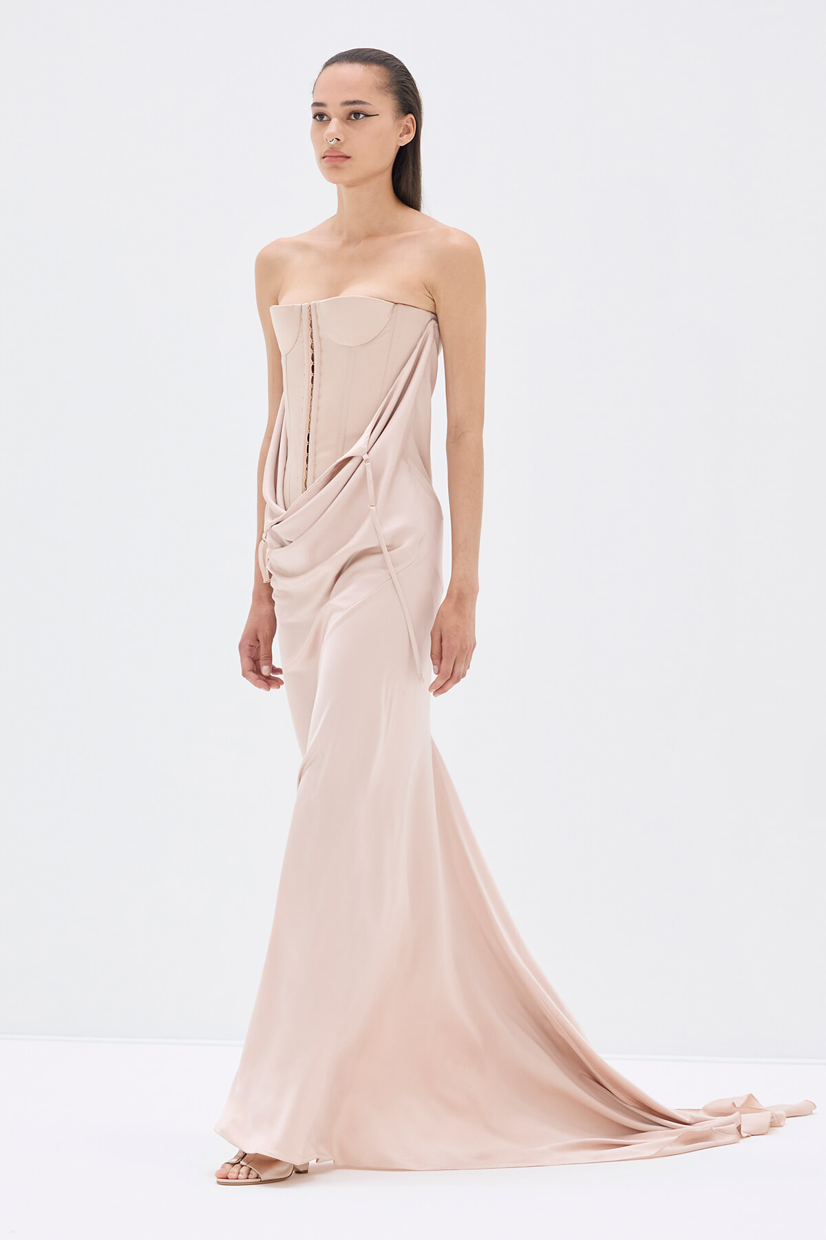 Model wearing a blush pink strapless gown with a corset-style bodice and draped sash, floor-length with a long train.