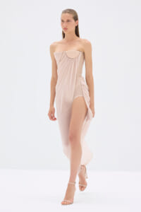 Model wearing a blush-pink strapless draped jumpsuit with a high-side slit and nude heeled sandals, standing against a white background.