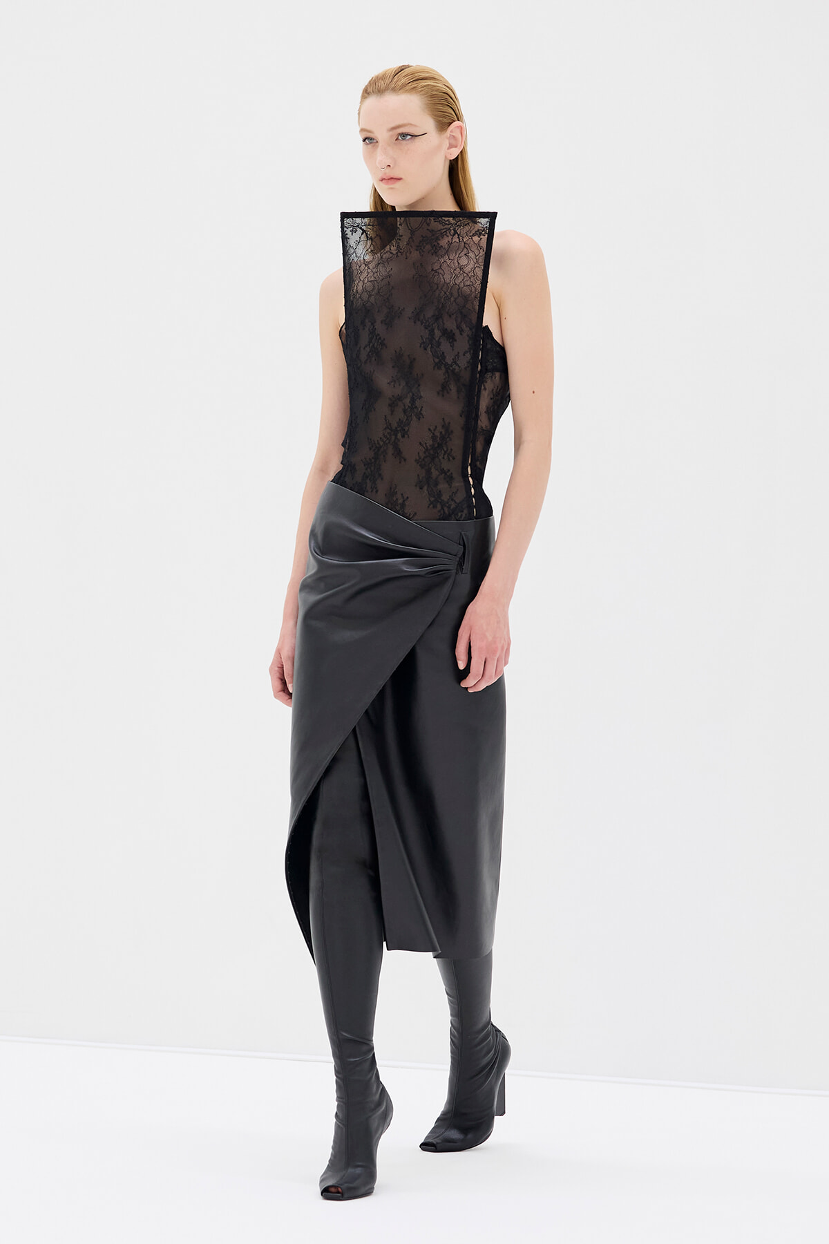 Model wearing a black lace high-neck sheer top with a draped black wrap skirt and black knee-high boots in a studio setting.