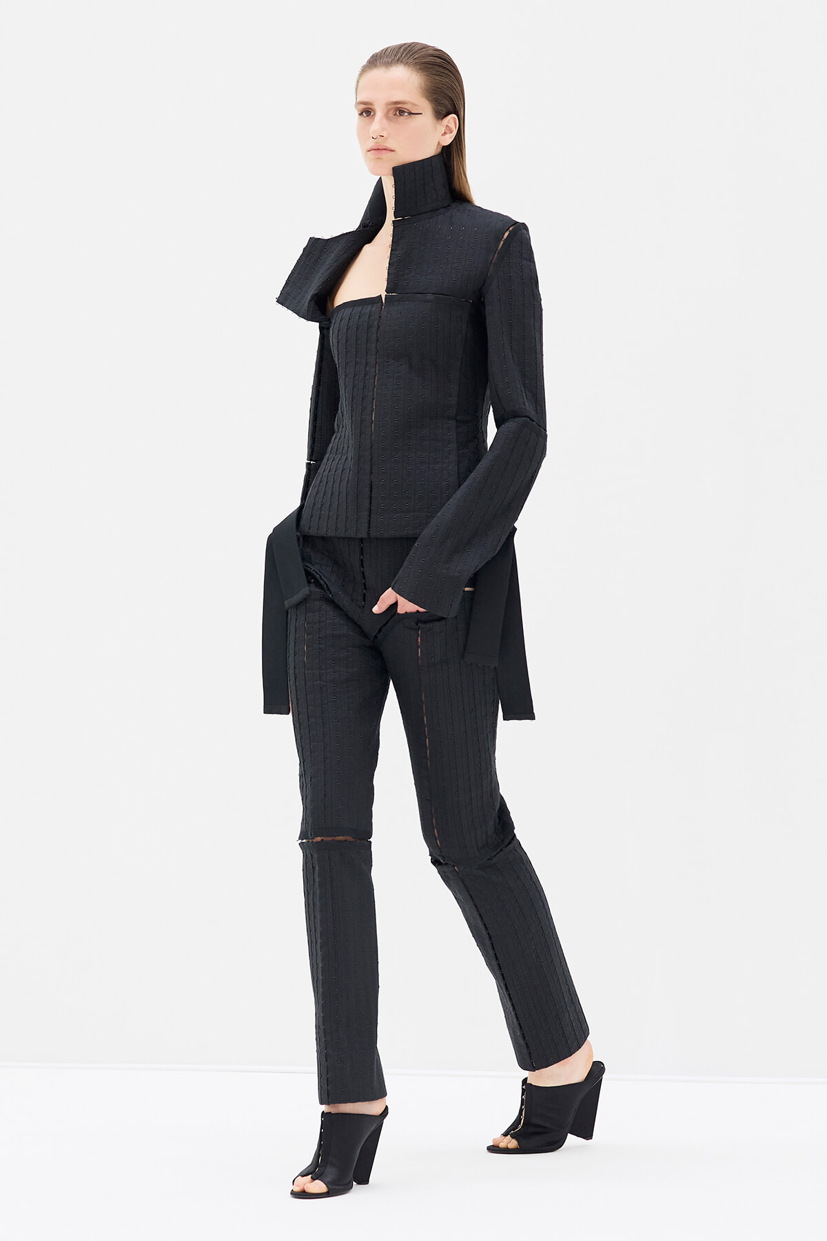 Model wearing a black textured two-piece suit with a high-collar jacket, fitted top, and cropped tapered pants; open-toe heeled sandals.