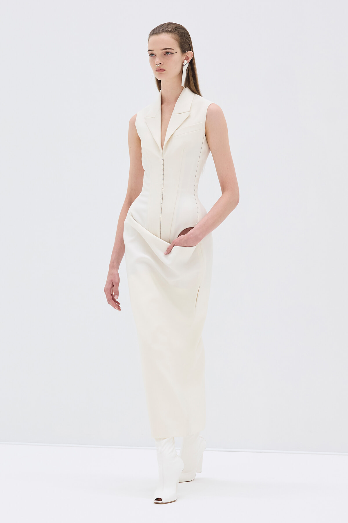 Ivory sleeveless dress with a plunging lapel collar, front zipper, and side pockets, paired with white knee-high heeled boots in a studio setting