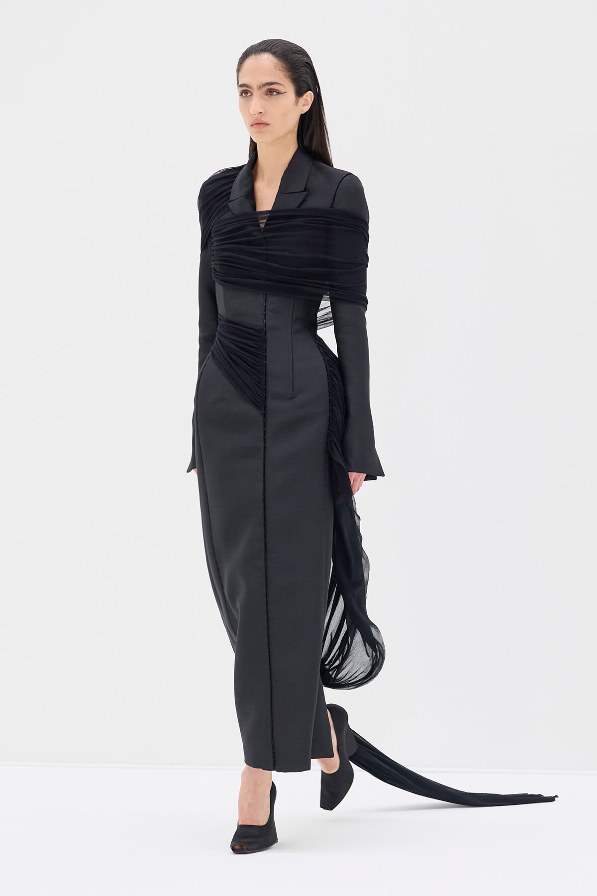 Full-length view of a model in a black sculptural gown with a wrapped bodice and long train extending behind her.