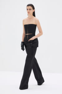 Model in a black strapless corset top and wide-leg pants, with a large tied bow at the waist and black gloves in a white studio setting