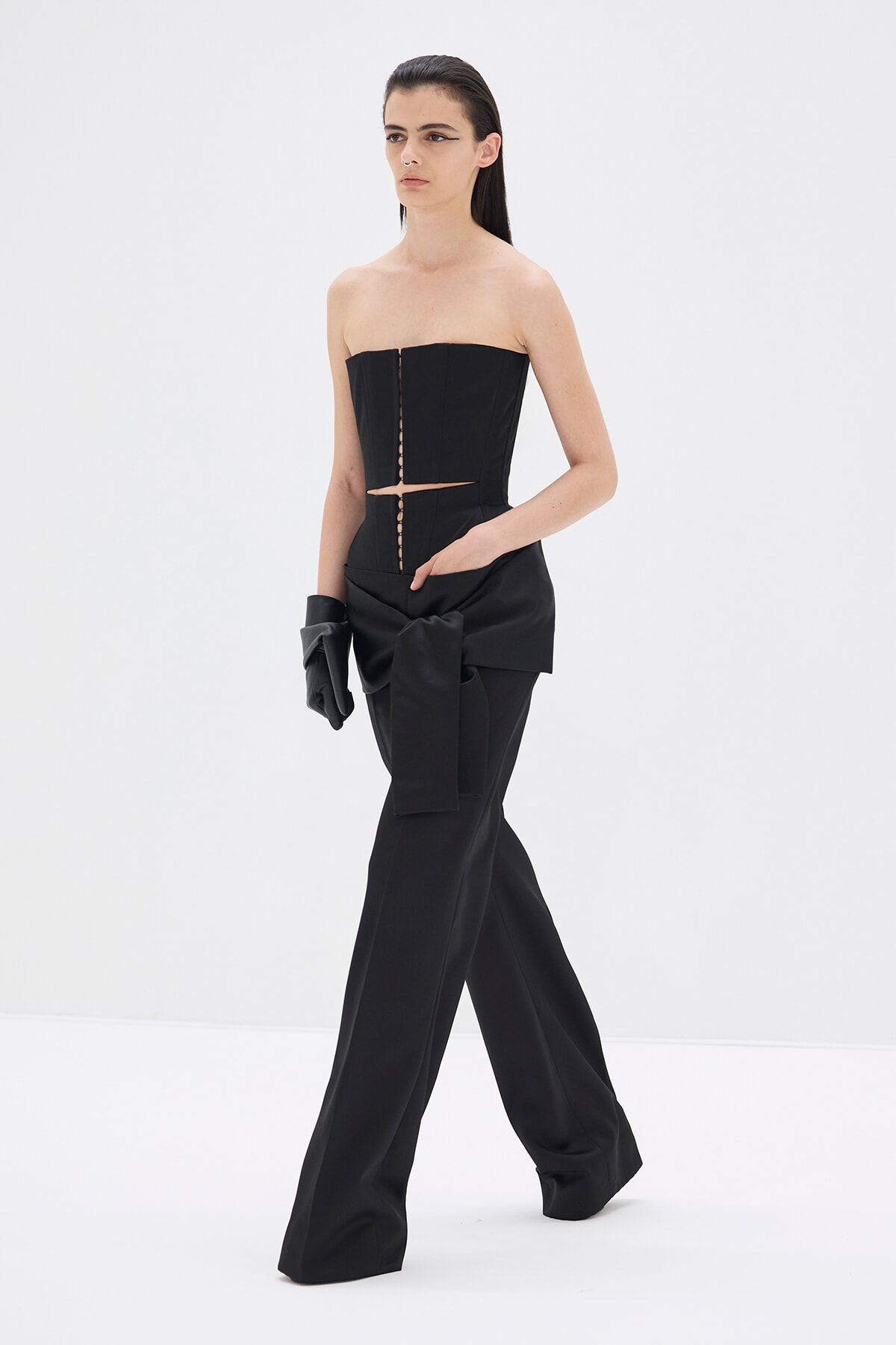 Model in a black strapless corset top and wide-leg pants, with a large tied bow at the waist and black gloves in a white studio setting