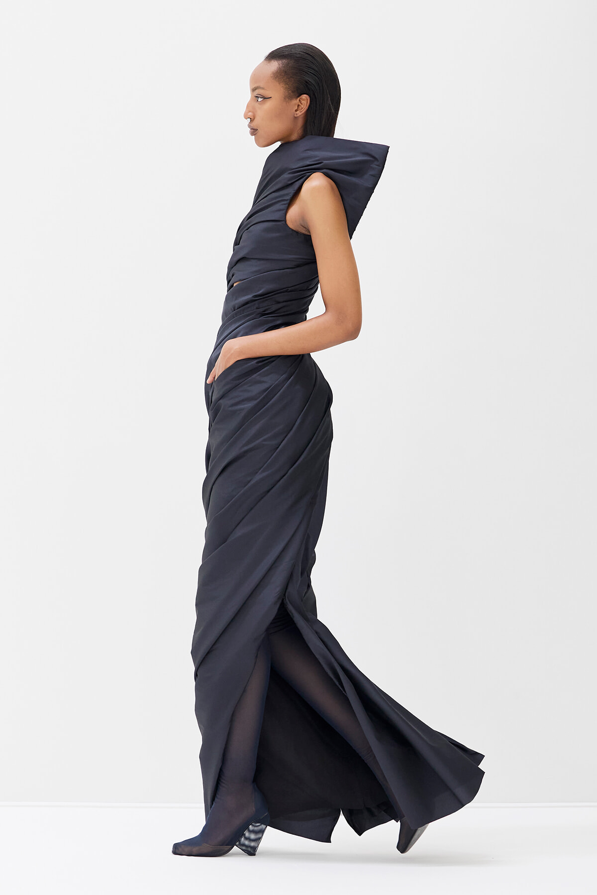 Side view of a woman in a dark navy draped gown with a sculptural collar, walking.