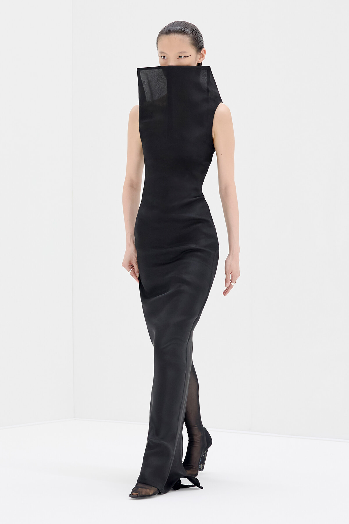 Full-length black sleeveless gown with a tall sculptural collar covering the lower face, worn by a model in a white studio.
