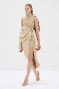 Model in a beige, high-neck sleeveless dress with a dramatic draped wrap and knee-high slit, wearing strappy sandals.