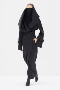 Model wearing an all-black, oversized coat with a large draped scarf that covers the lower face, standing in a white studio.