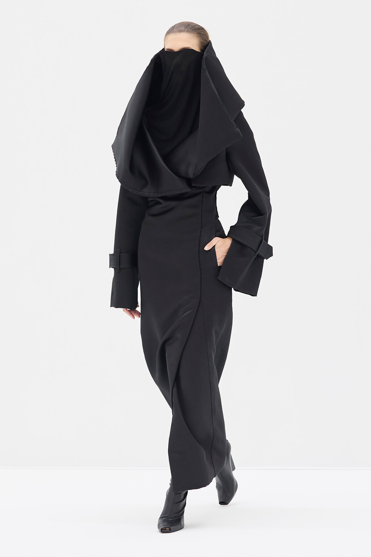 Model wearing an all-black, oversized coat with a large draped scarf that covers the lower face, standing in a white studio.