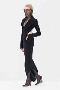Model in a black tailored wrap dress with a side cutout, lace veil over the face, standing in sheer tights and patterned heeled shoes.