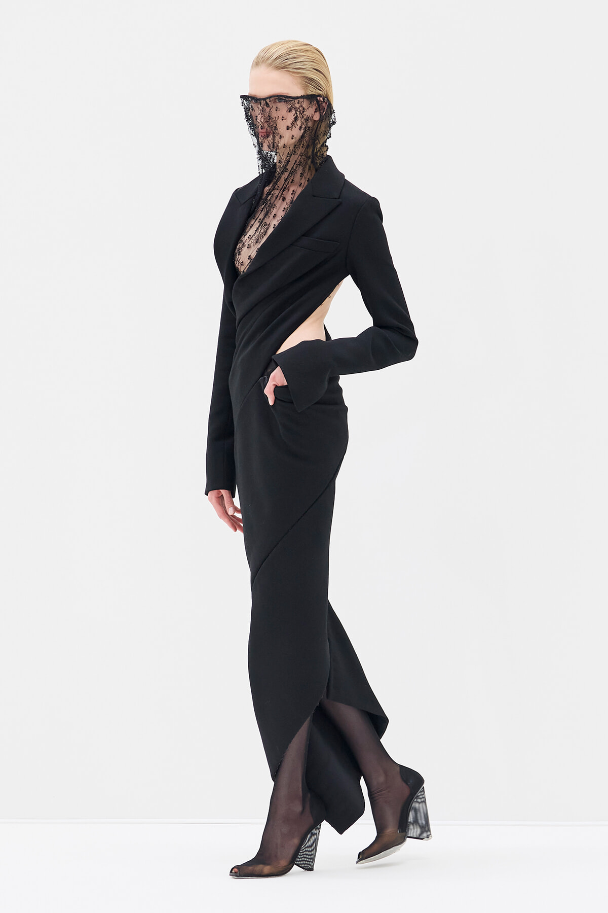 Model in a black tailored wrap dress with a side cutout, lace veil over the face, standing in sheer tights and patterned heeled shoes.