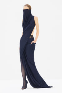 Model wearing a navy blue draped gown with a high neck covering the lower face, side cutouts, and a long trailing train.