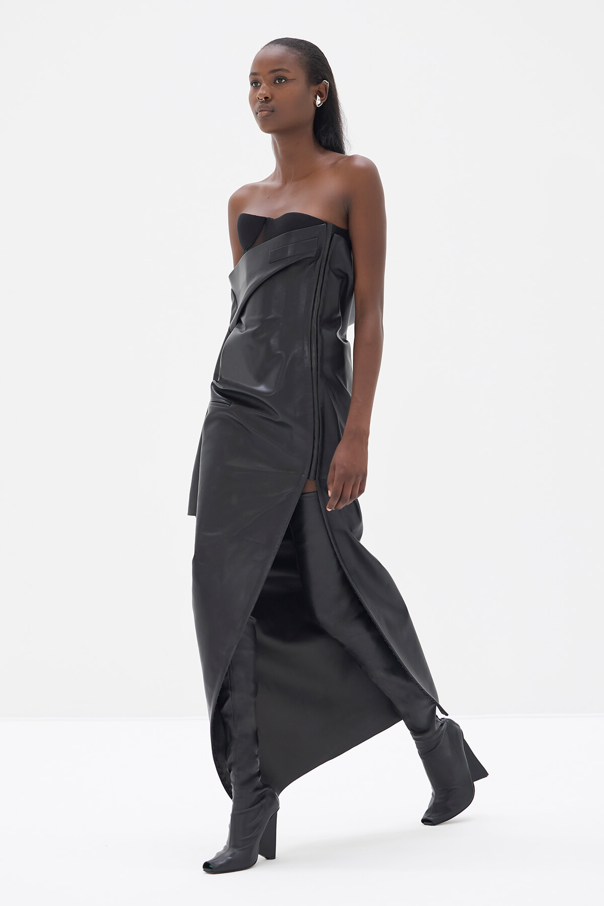 Model wearing a strapless black leather dress with an asymmetrical wrap and matching knee-high boots in a studio setting.