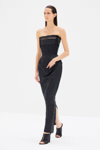 Model wearing a black strapless corset-style jumpsuit with structured bodice and front seams, paired with black heeled sandals against a white studio background.
