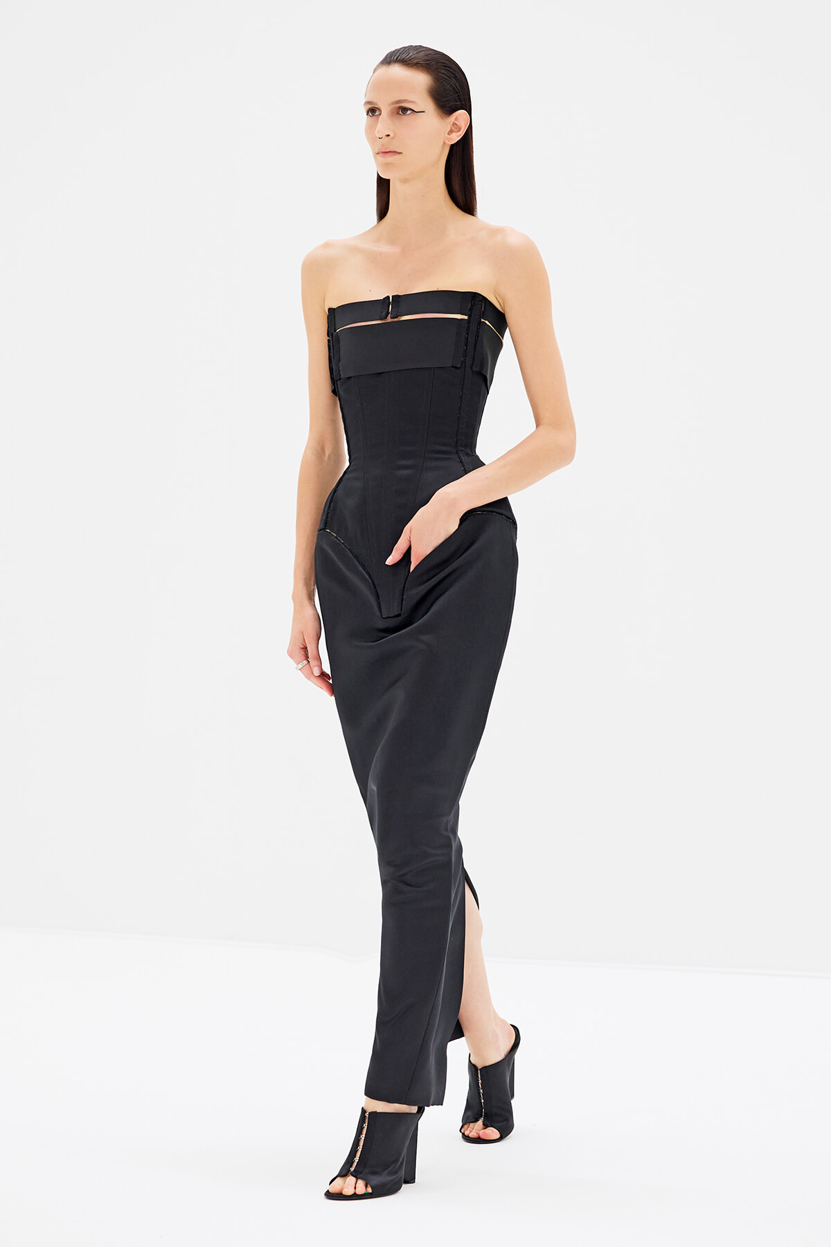 Model wearing a black strapless corset-style jumpsuit with structured bodice and front seams, paired with black heeled sandals against a white studio background.