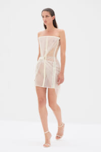 Model wearing a sheer cream strapless mini dress with a front waist knot and white ankle-strap heels, posing in a studio.