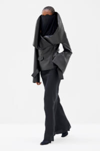 Person wearing a large black leather-like coat with a dramatic draped collar that covers the lower face, paired with black wide-leg pants.