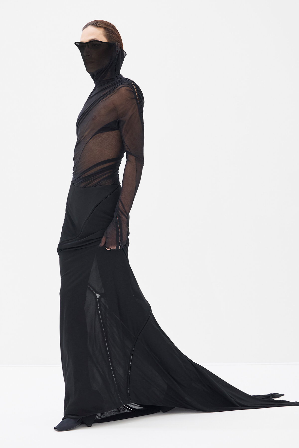 Model in a sheer black mesh top and long black gown with a sweeping train, posing against a white studio background.